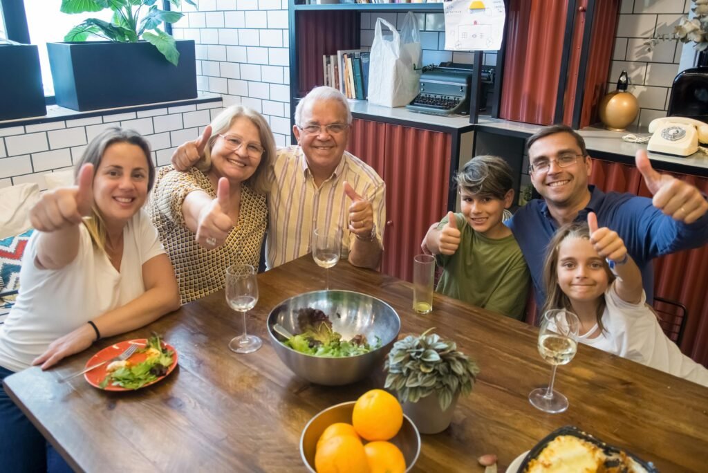 "Idosa sorrindo ao redor da mesa com sua família reunida, compartilhando um almoço em um momento de união e alegria."